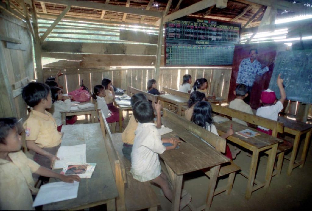 A candid photo of a classroom shows a dozen young Indonesian children seated at simple wooden desks and benches inside a rustic building with slatted walls and a wooden ceiling. A male teacher in a patterned shirt stands at the front, pointing to a blackboard while one student raises their hand.