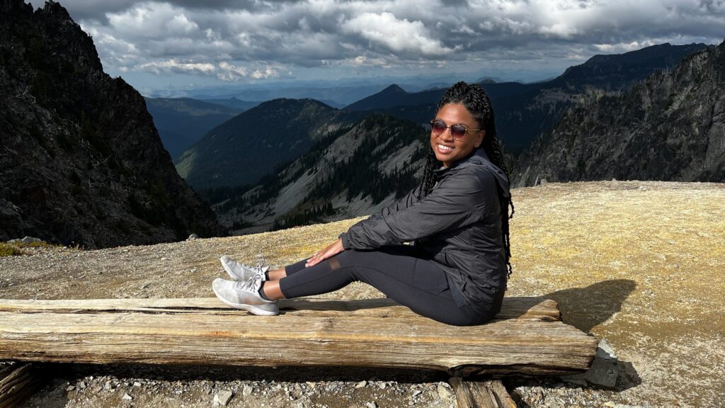 A woman sitting on a log high up on a rocky surface, with mountains in the background
