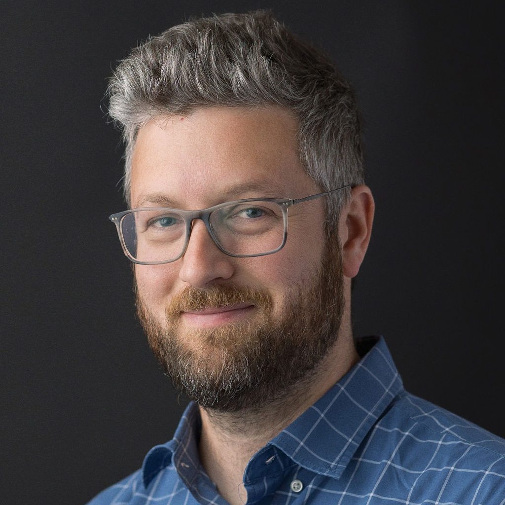 A photograph of a bearded man wearing glasses and a blue button-up shirt, set against a black background.