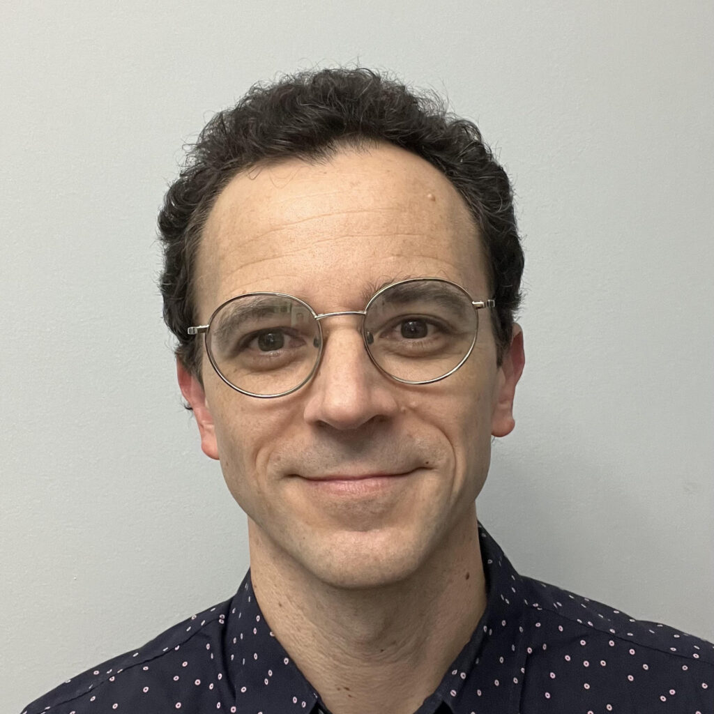 A man with rounded glasses and a button-up shirt smiles slightly, in front of an off-white background.